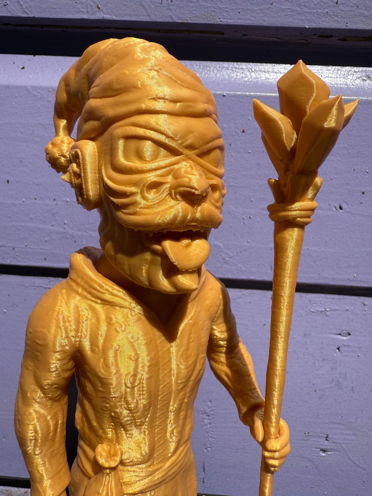 Gold-toned Galaktic Gang 8” art toy with a hat, staff, exaggerated facial features and long tongue, displayed against a purple wooden background.