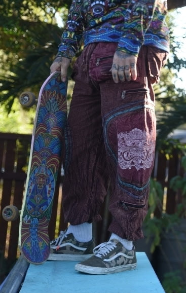 Model wearing handmade Chris Dyer Dyer-Jawnz parachute pants in burgundy with patchwork details while holding a colorful skateboard.