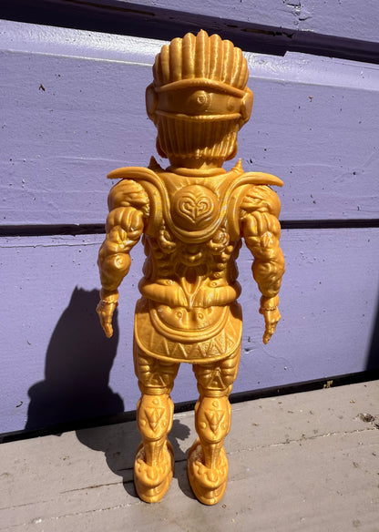 The Galaktic Dude Figurine, an 8-inch art toy in a vibrant yellow armored suit with intricate molded details, stands upright facing away on a sunlit surface before a purple wooden wall.
