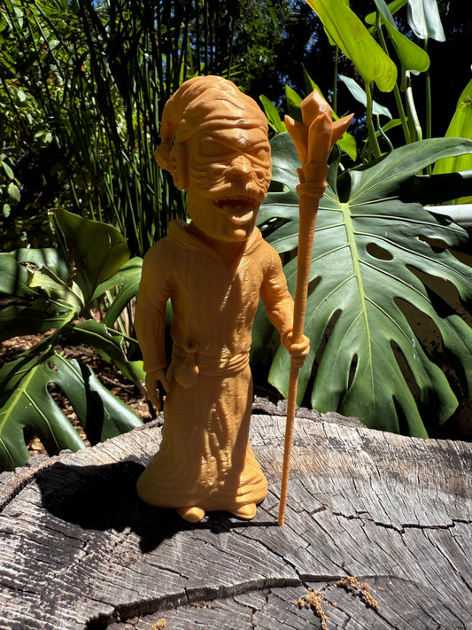 Wacky Wizard 3D print golden figurine of a robed figure holding a staff, displayed outdoors on a weathered log with lush green tropical foliage in the background.
