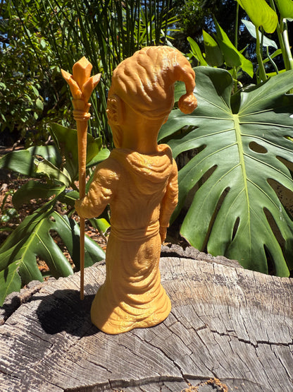 Wacky Wizard 3D print golden figurine of a robed Wizard holding a staff, photographed outdoors on a weathered log with large green tropical leaves in the background.