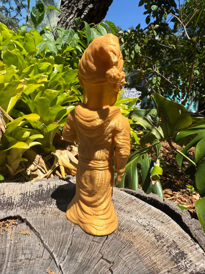 Wacky Wizard 3D print golden-yellow figurine of a robed Wizard figure with a tall sculpted head, standing on a weathered wooden log outdoors with green foliage and trees in the background.
