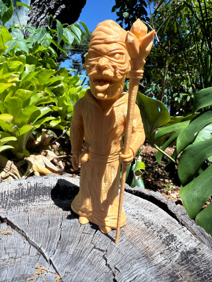 Wacky Wizard 3D print golden figurine of a robed character holding a Crystal staff, displayed outdoors on a weathered tree stump with lush green foliage in the background.