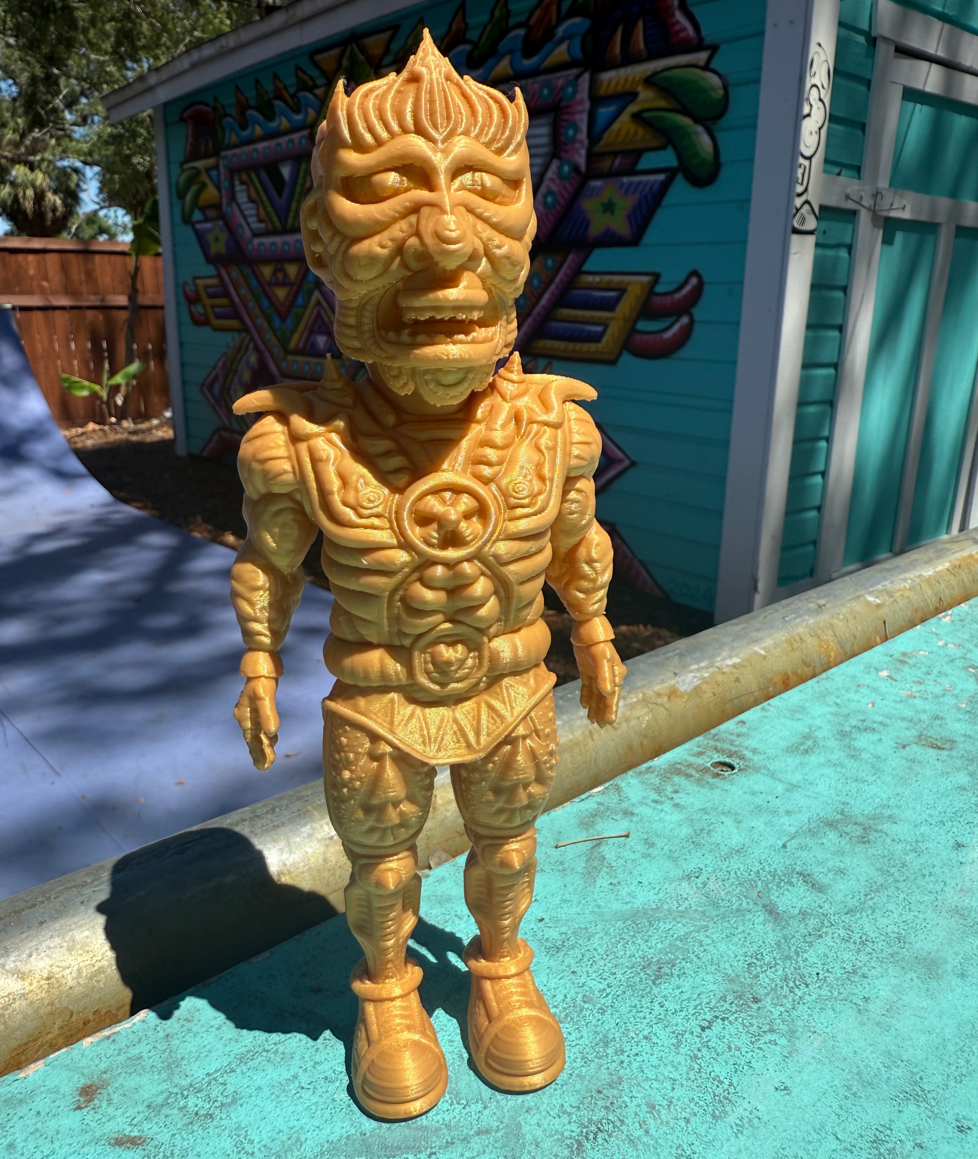The Galaktic Dude Figurine, a golden warrior figurine with a tribal mask head stands on a turquoise painted surface outdoors in front of a colorful building.  