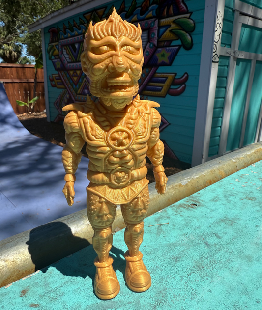 The Galaktic Dude Figurine, a golden warrior figurine with a tribal mask head stands on a turquoise painted surface outdoors in front of a colorful building.  