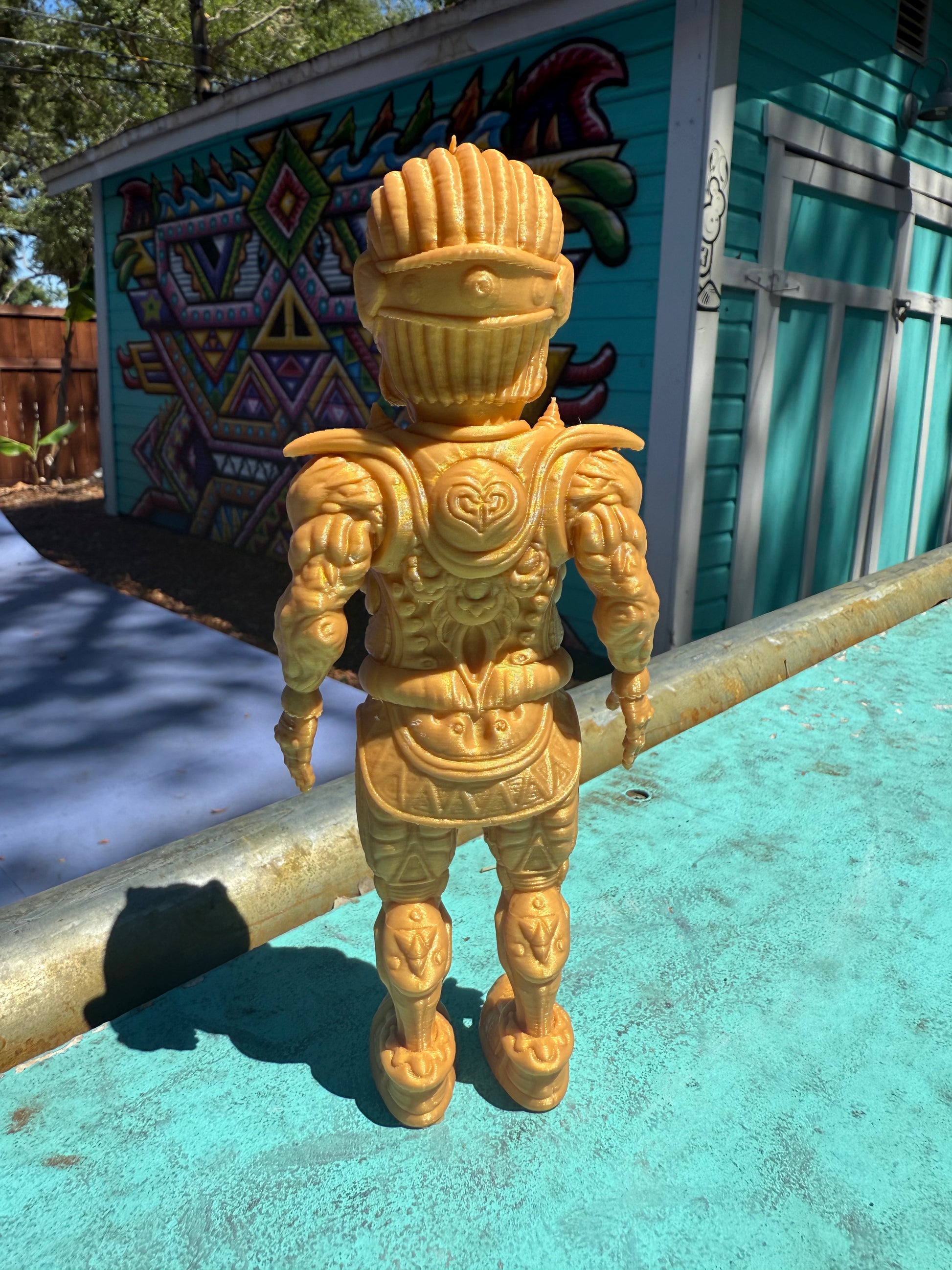 The Galaktic Dude Figurine, a golden warrior figurine with a tribal mask head stands on a turquoise painted surface outdoors in front of a colorful building.  