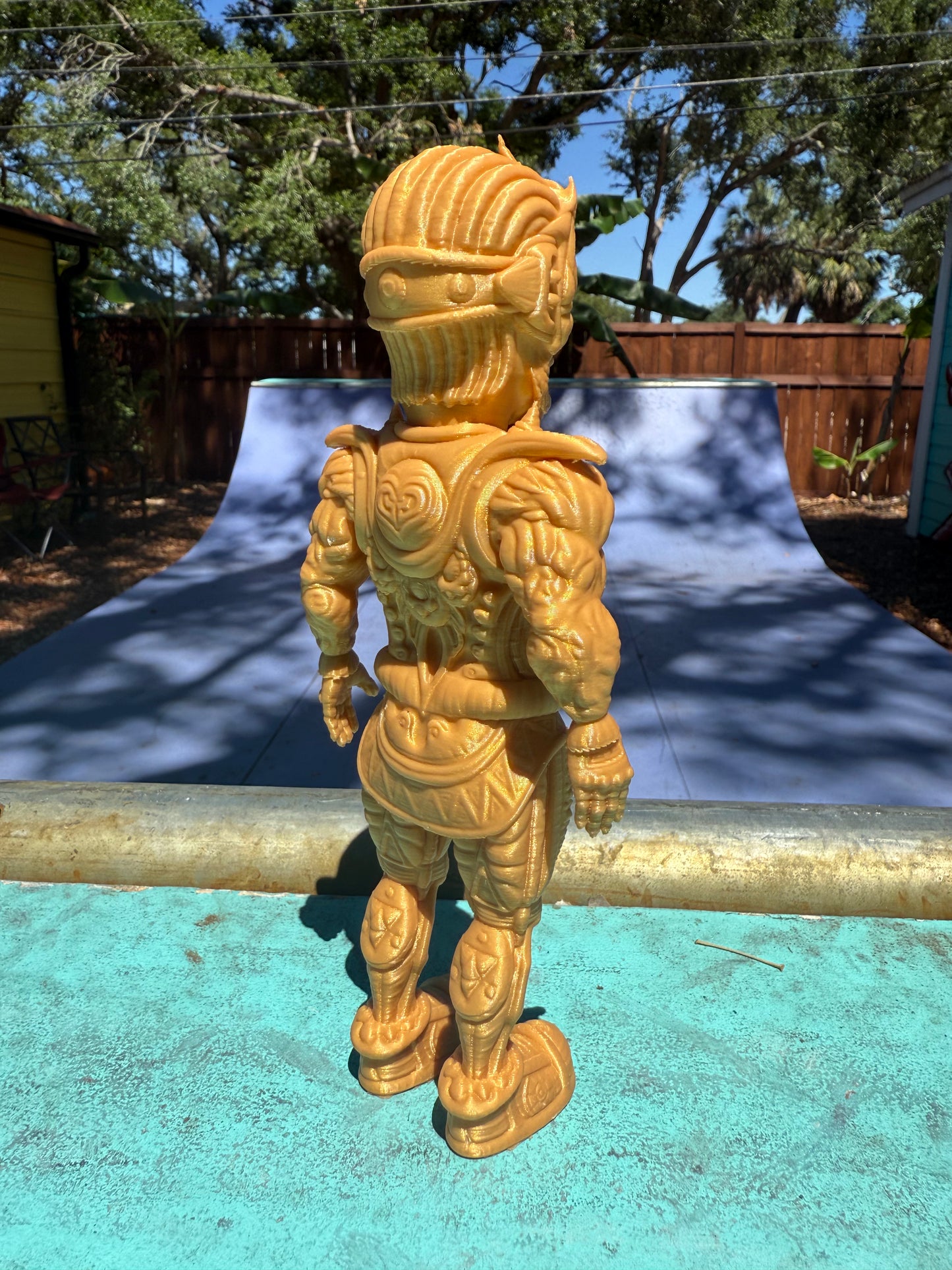 The Galaktic Dude Figurine, a golden warrior figurine with a tribal mask head stands on a turquoise painted surface outdoors in front of a colorful building.  