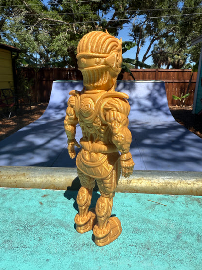 The Galaktic Dude Figurine, a golden warrior figurine with a tribal mask head stands on a turquoise painted surface outdoors in front of a colorful building.  