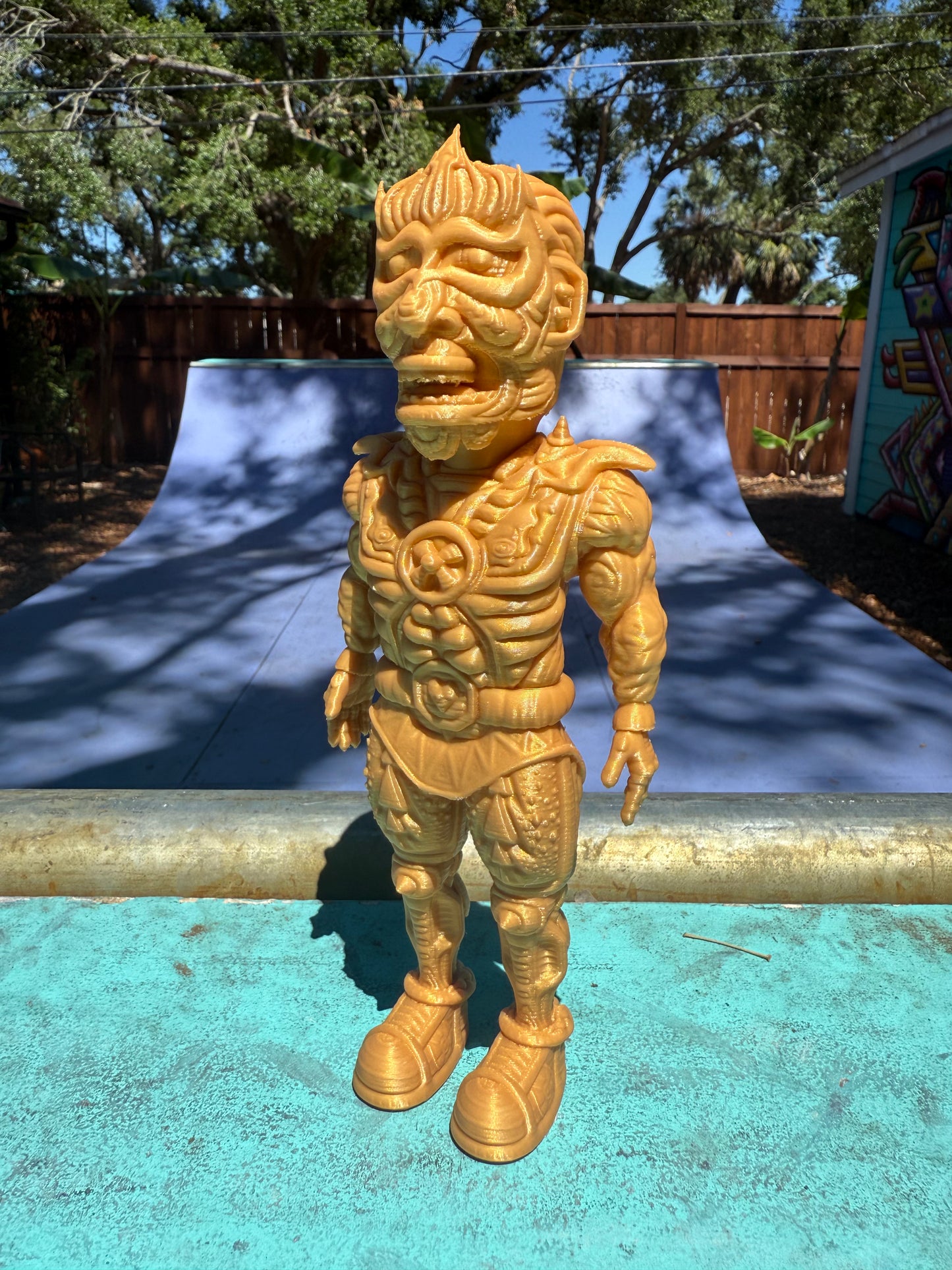 The Galaktic Dude Figurine, a golden warrior figurine with a tribal mask head stands on a turquoise painted surface outdoors in front of a colorful building.  
