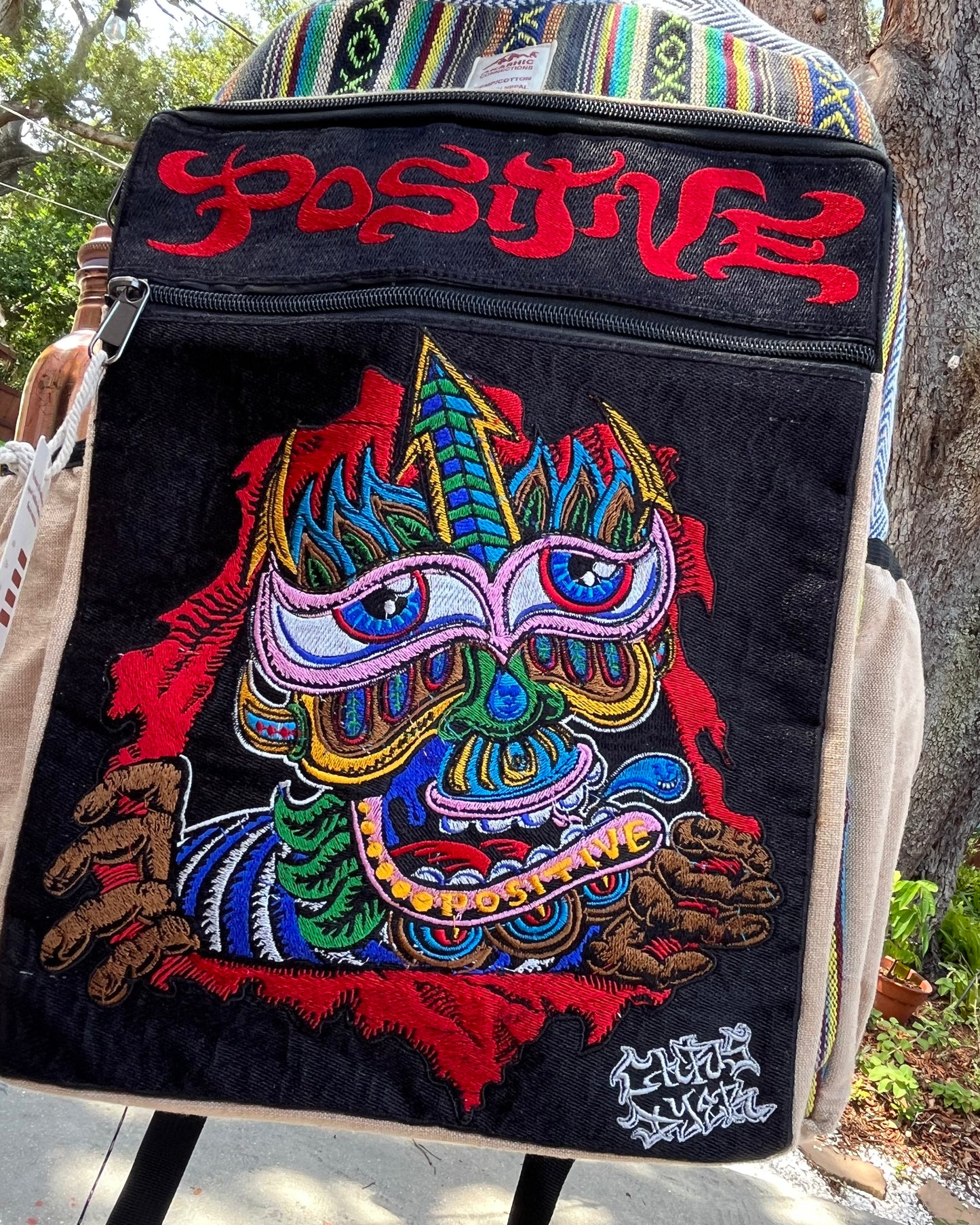 Akashic Connections × Chris Dyer “Positive” hemp backpack outdoors, featuring a psychedelic, colorful mask design and embroidered “POSITIVE” text on a sunny day.