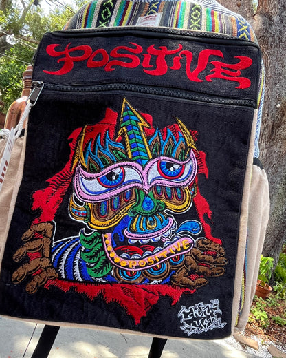 Akashic Connections × Chris Dyer “Positive” hemp backpack outdoors, featuring a psychedelic, colorful mask design and embroidered “POSITIVE” text on a sunny day.