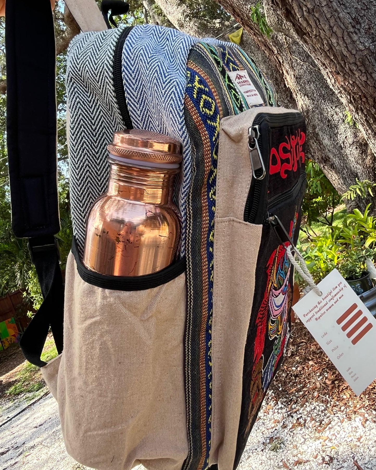 The Akashic Connections × Chris Dyer “Positive” hemp backpack outdoors with a copper bottle in its side pocket and a color chart tag, set against lush green trees.
