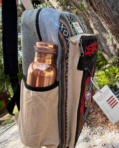 The Akashic Connections × Chris Dyer “Positive” hemp backpack outdoors with a copper bottle in its side pocket and a color chart tag, set against lush green trees.