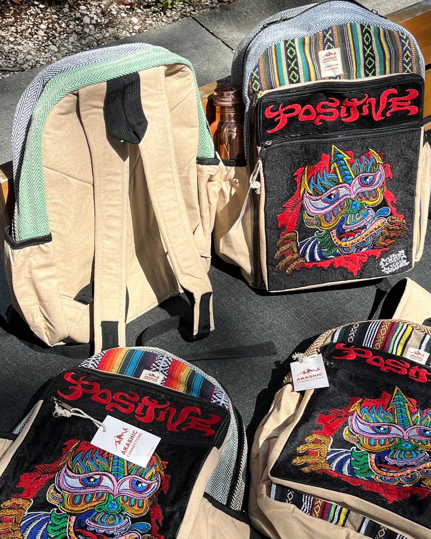 Four “Positive” hemp backpacks by Chris Dyer × Akashic Connections with vivid psychedelic embroidery, unique patterns, neutral fabrics, zippers and side pockets—one holding a water bottle—rest on sunlit ground.