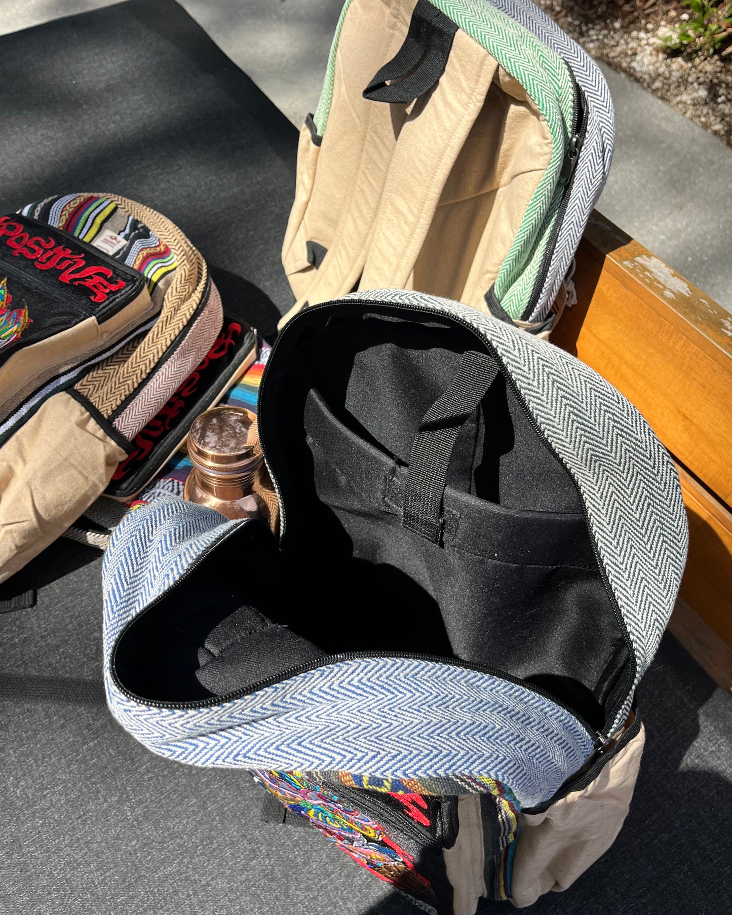 Two colorful Akashic Connections × Chris Dyer “Positive” hemp backpacks with striped tribal designs rest on an outdoor bench; one contains a copper water bottle, the other is empty. Strong sunlight creates sharp shadows on the ground.