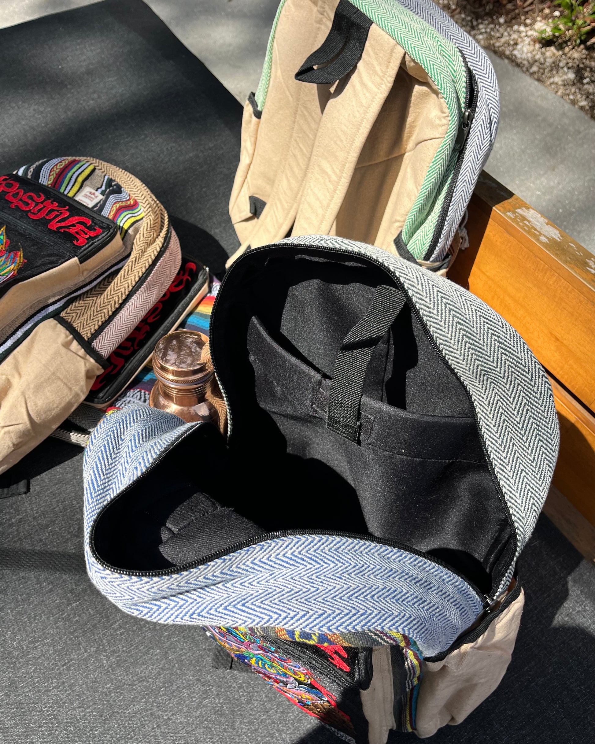 Two colorful Akashic Connections × Chris Dyer “Positive” hemp backpacks with striped tribal designs rest on an outdoor bench; one contains a copper water bottle, the other is empty. Strong sunlight creates sharp shadows on the ground.