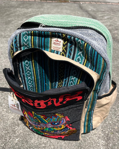 “Positive” Hemp Backpack by Chris Dyer and Akashic Connections unzipped on sunlit concrete, featuring colorful psychedelic stripes, a tan fabric base, and a vibrant tribal-style graphic front pocket.