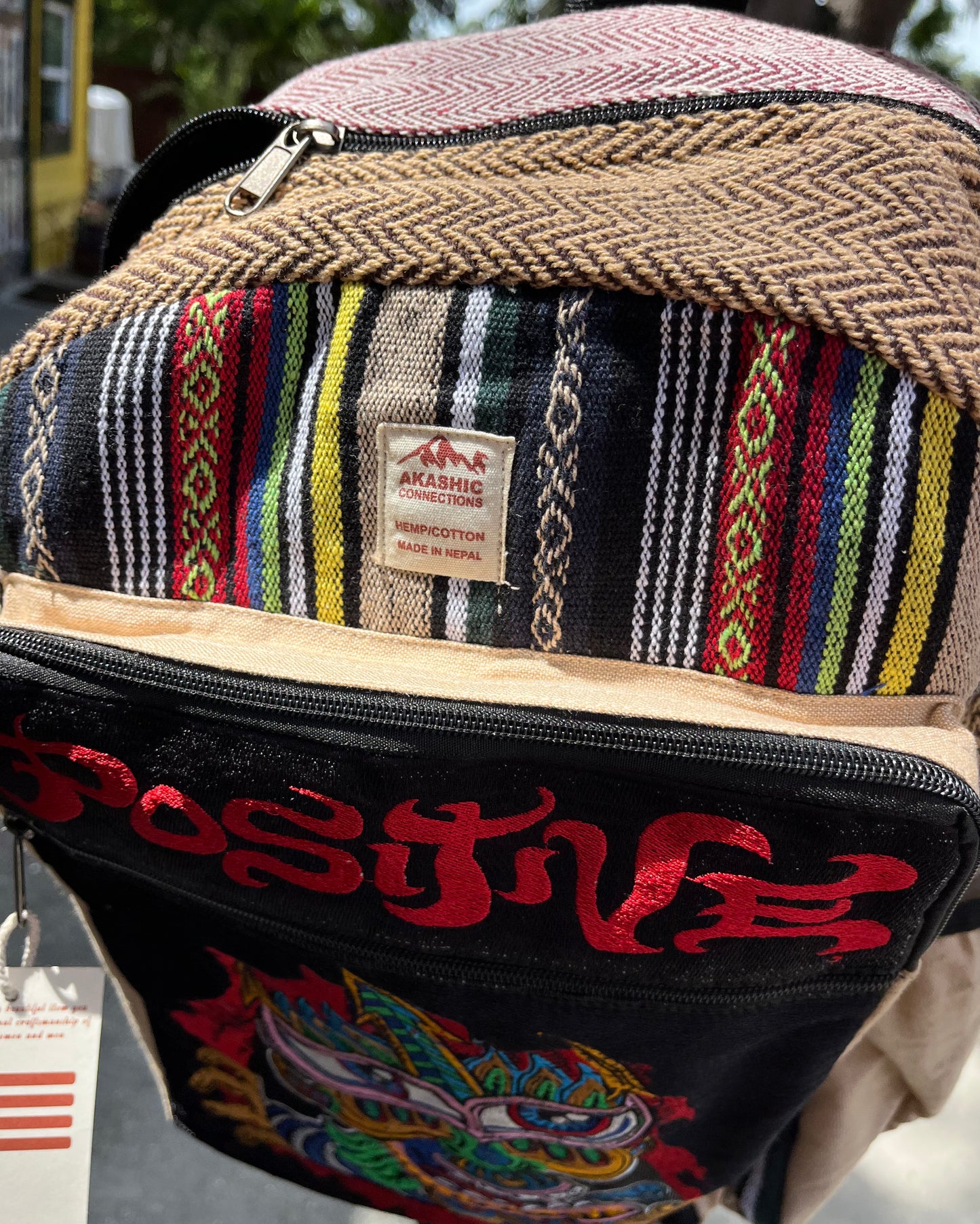 Akashic Connections × Chris Dyer “Positive” hemp backpack with woven multicolor stripes, a front pocket showing red embroidered text and patch, plus a small fabric label reading “Akashic Connection - Made in Nepal.”.