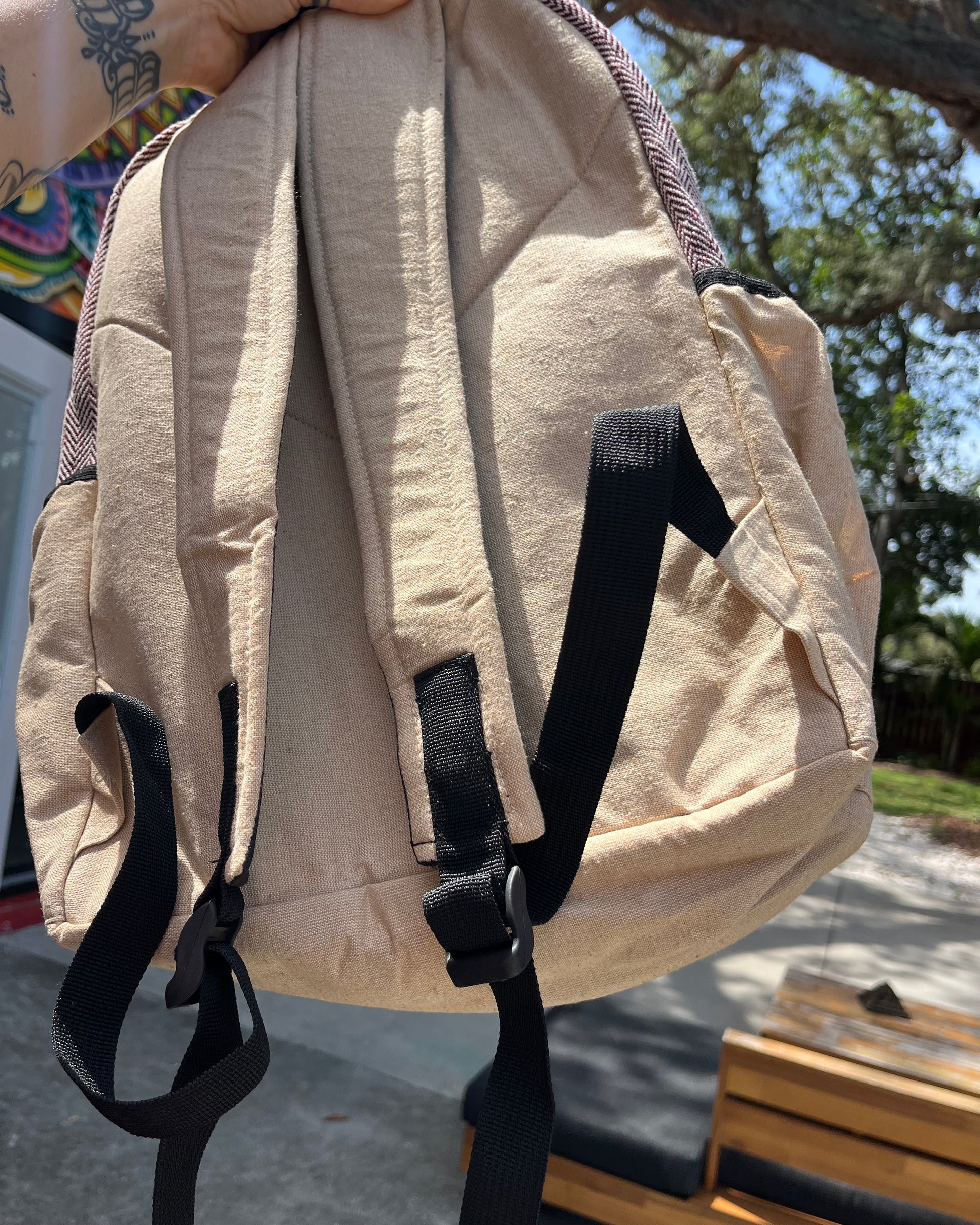 Hand holding the Akashic Connections × Chris Dyer “Positive” Hemp Backpack outdoors, with a psychedelic, colorful mural in sunlight behind, plus a wooden bench and tree branches casting shadows.