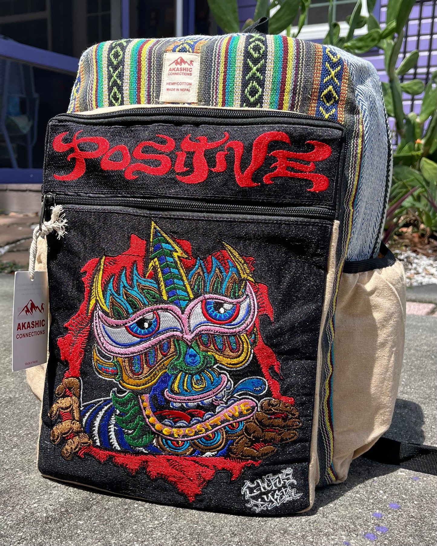 Akashic Connections × Chris Dyer “Positive” hemp backpack with striped fabric, a colorful psychedelic monster patch, red “POSITIVE” text, and AKASHIC CONNECTION tag, displayed outdoors on concrete.
