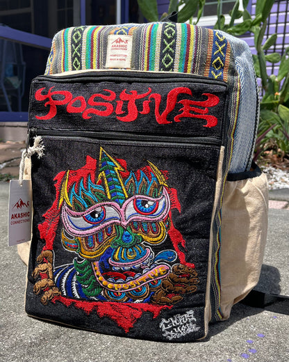 Akashic Connections × Chris Dyer “Positive” hemp backpack with striped fabric, a colorful psychedelic monster patch, red “POSITIVE” text, and AKASHIC CONNECTION tag, displayed outdoors on concrete.