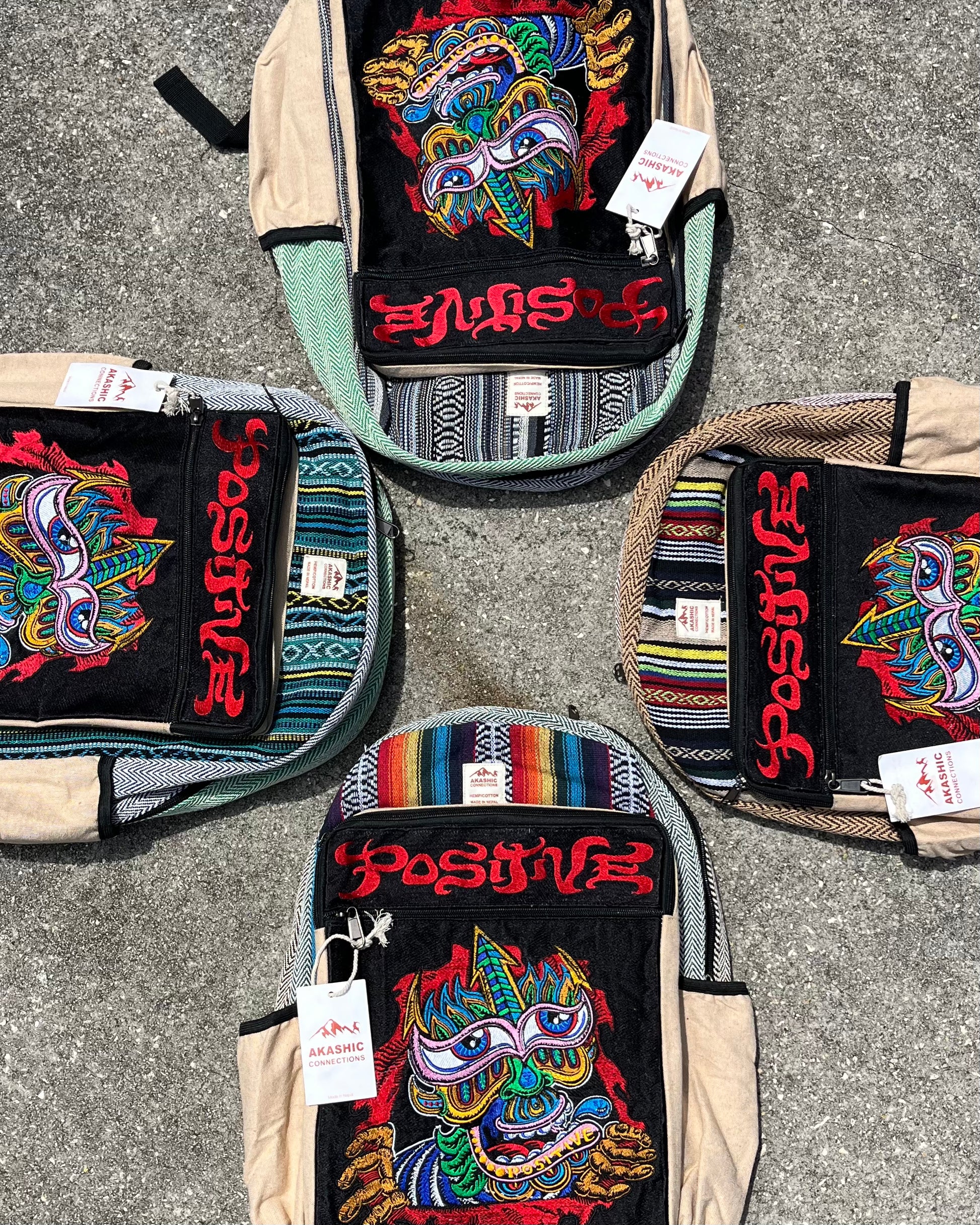Four colorful Akashic Connections × Chris Dyer “Positive” hemp backpacks with psychedelic monster faces and striped patterns, each tagged and embroidered with “positive” in red, arranged in a circle on concrete.