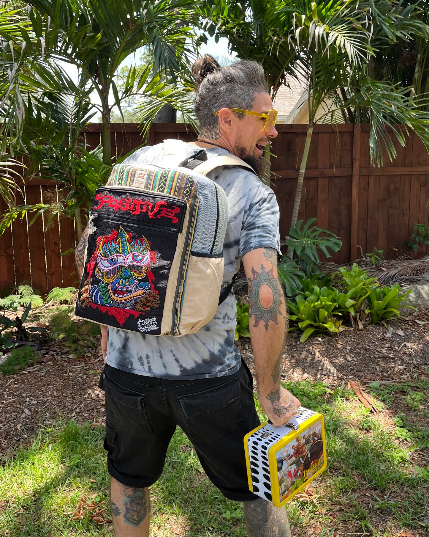 Tattooed man with beard and topknot wearing yellow sunglasses, tie-dye shirt, black cargo shorts, and the colorful Akashic Connections × Chris Dyer “Positive” Hemp Backpack stands in a backyard lined with palm trees.