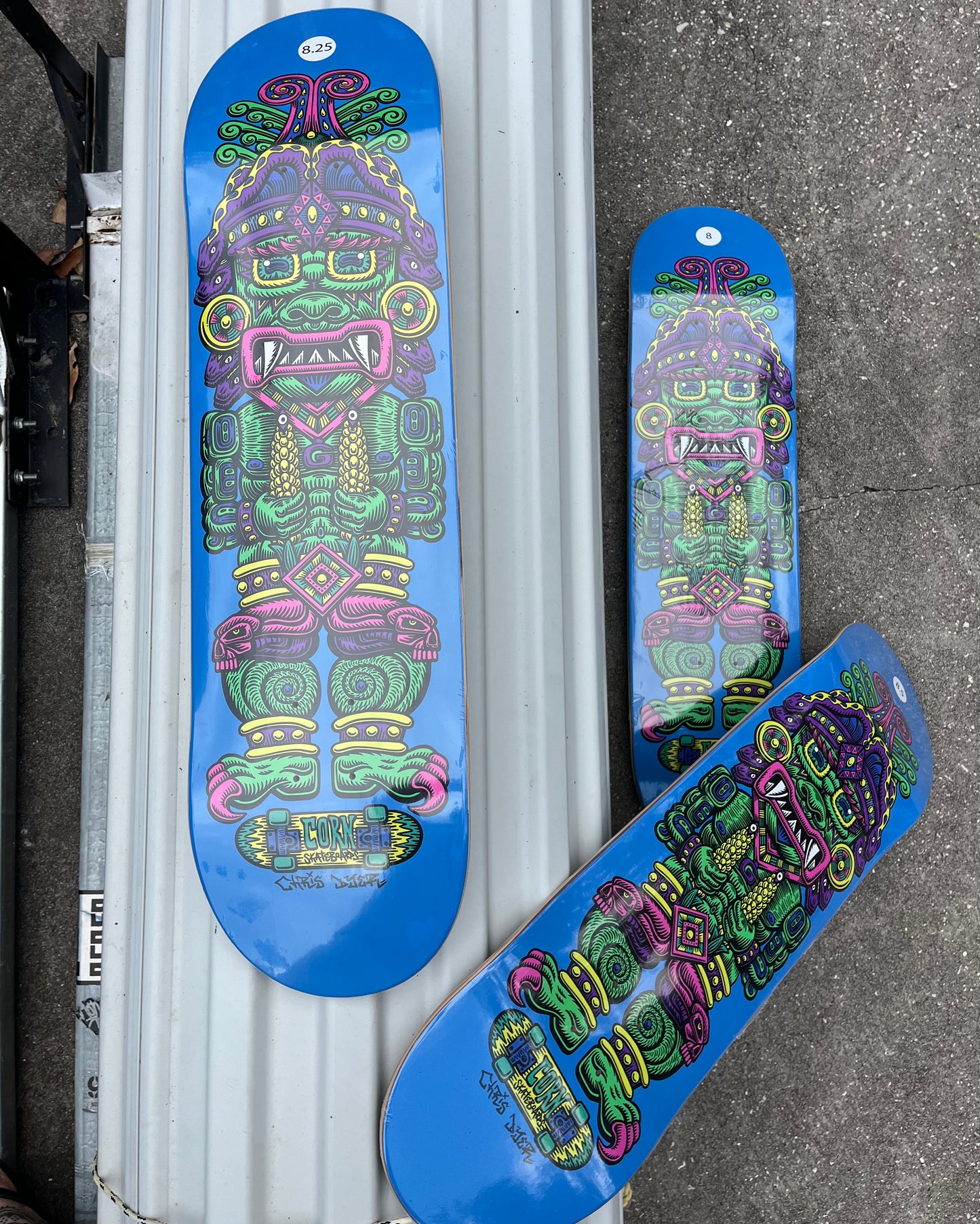 Three skateboard decks with a vibrant blue background and a colorful Aztec-style warrior graphic, displayed on a white corrugated metal surface and concrete ground.