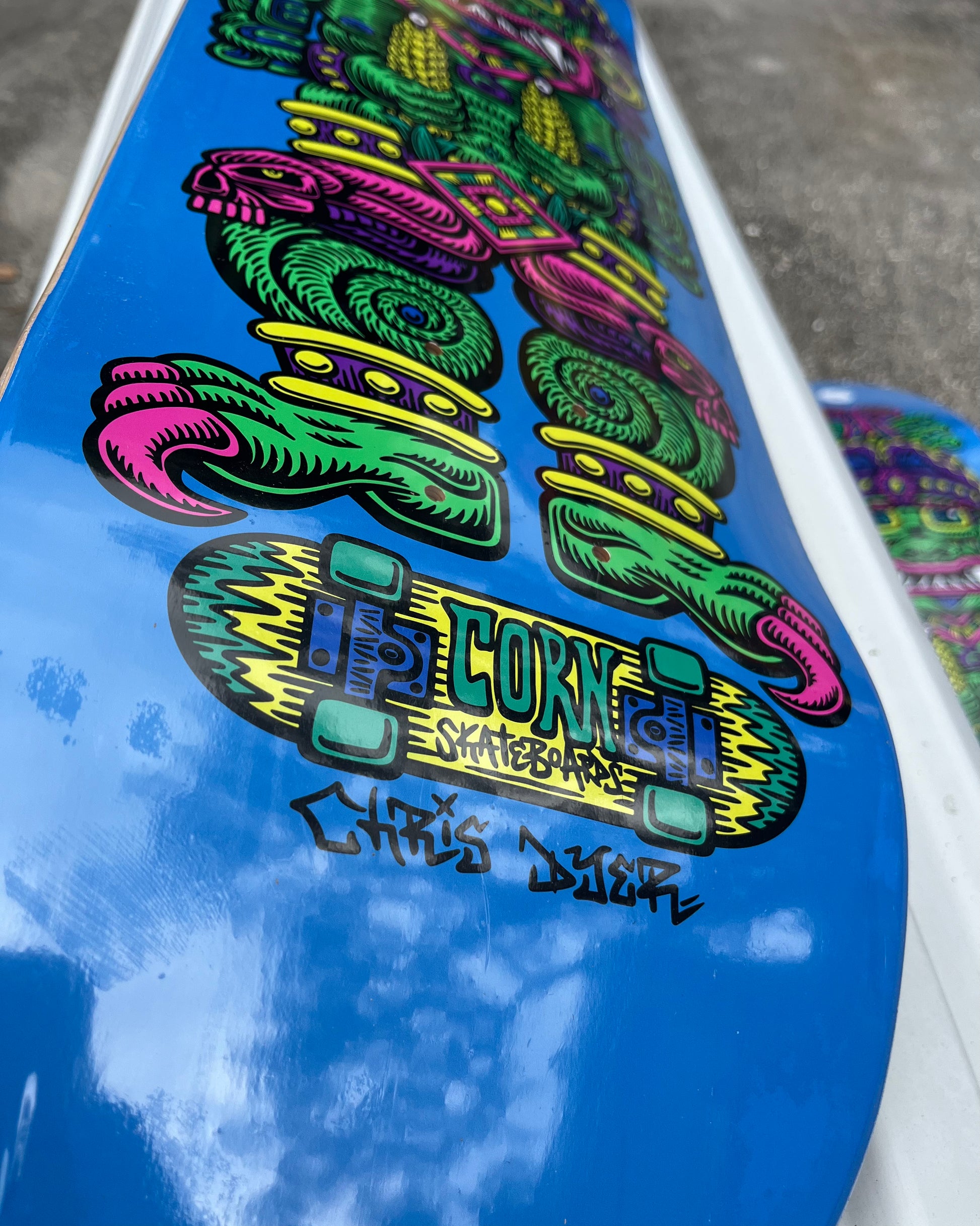 A skateboard deck with a vibrant blue background featuring Chris Dyer’s colorful psychedelic depiction of Wiracocha, the Incan Corn God, in green, purple, and pink.