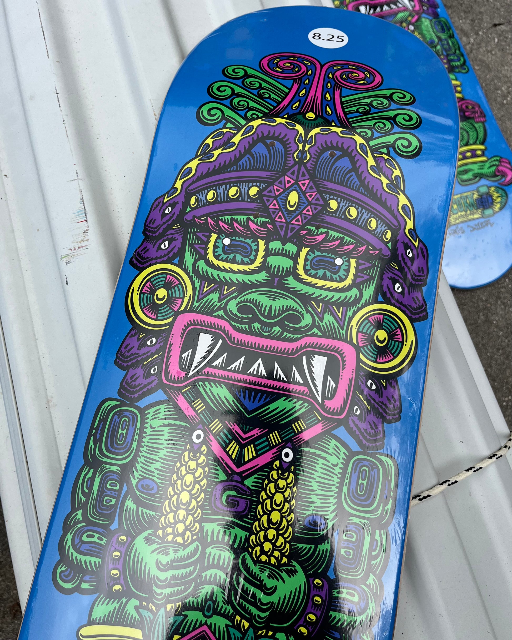 A skateboard deck with a vibrant blue background featuring Chris Dyer’s colorful psychedelic depiction of Wiracocha, the Incan Corn God, in green, purple, and pink.