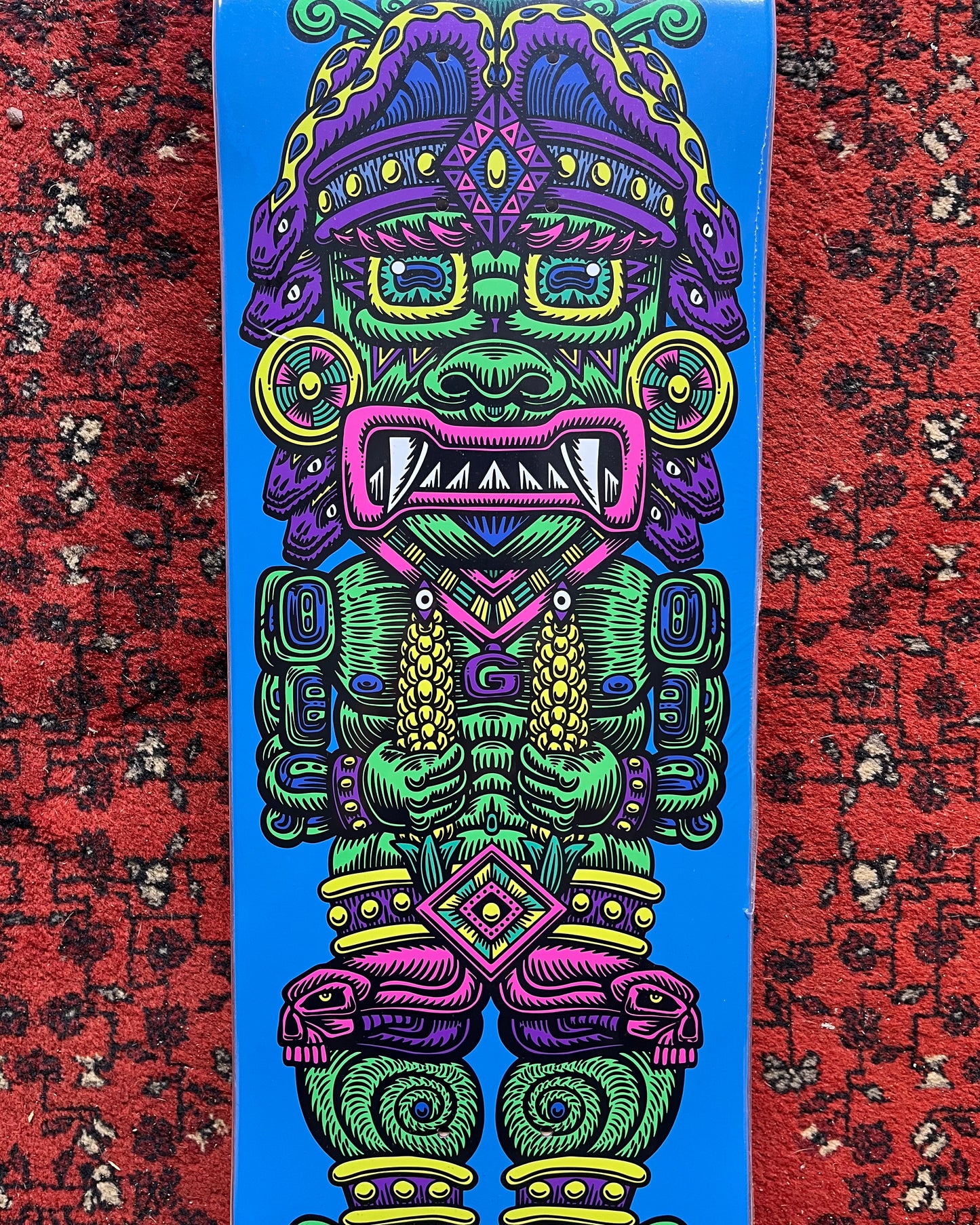 A skateboard deck featuring a highly detailed, colorful Aztec warrior graphic on a bright blue background, photographed standing upright on a red patterned rug.