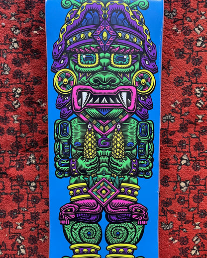 A skateboard deck featuring a highly detailed, colorful Aztec warrior graphic on a bright blue background, photographed standing upright on a red patterned rug.