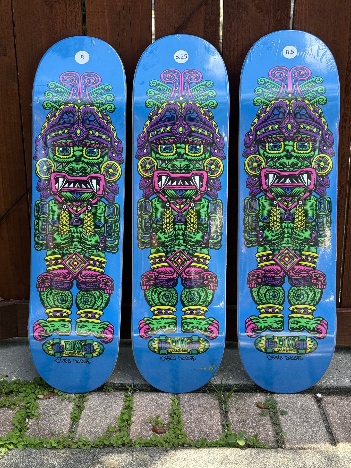 Three blue skateboard decks with colorful Aztec-style warrior graphics, standing upright against a dark wooden fence on a paved surface.