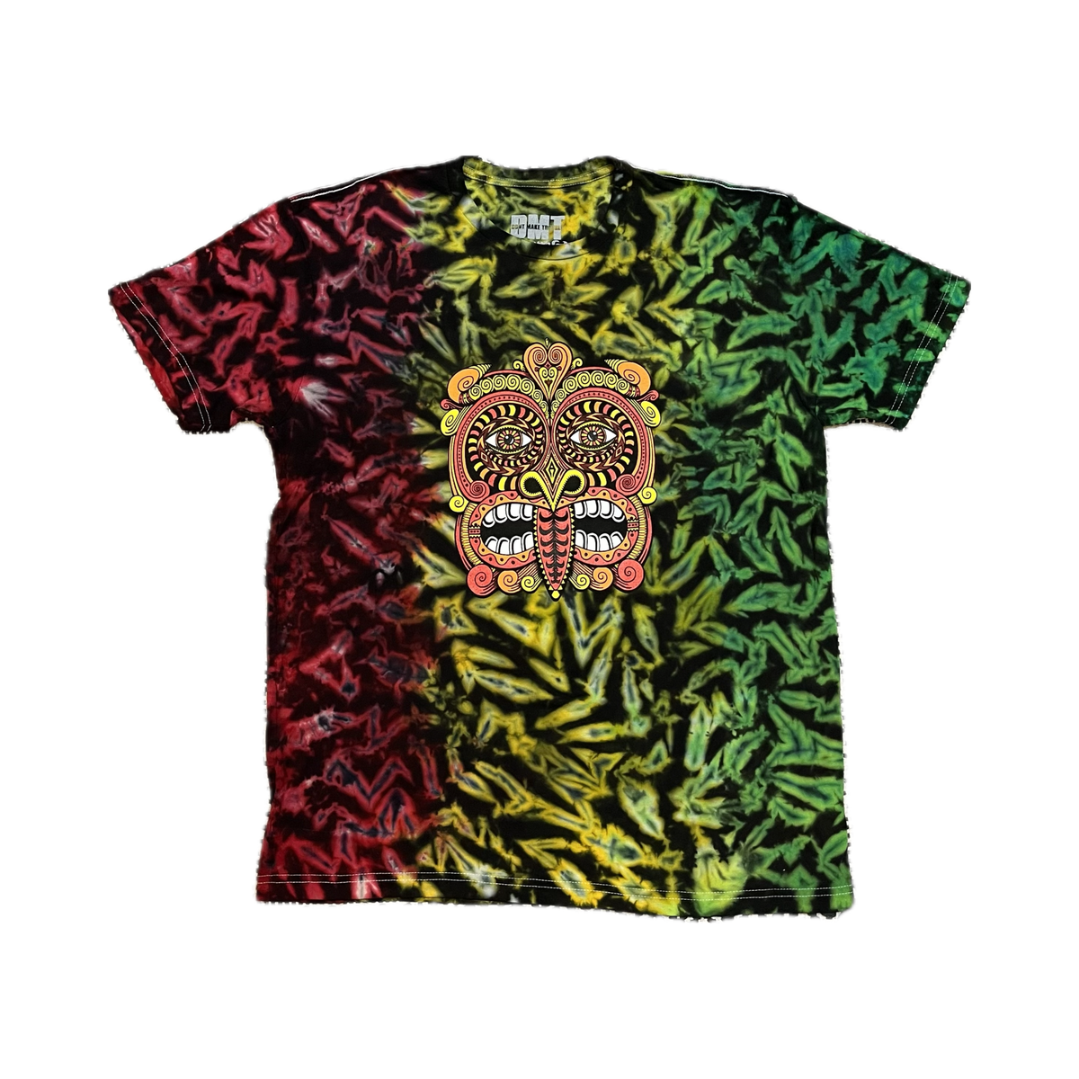 The Rainbow/Rasta Tribal Mask 2023 Tie Dye T Shirt with bold red, yellow, and green tie-dye and a large psychedelic Maori mask design lies flat on a black-and-white patterned rug.